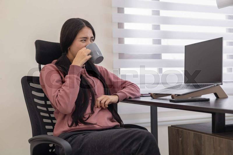 In a bright office, a young woman sits at her desk holding a cup of coffee, working