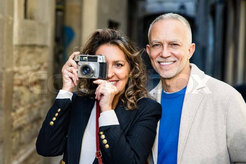 Mid adult tourist couple taking picture on vintage camera looking at camera. Senior woman taking photography in a city street with her husband.
