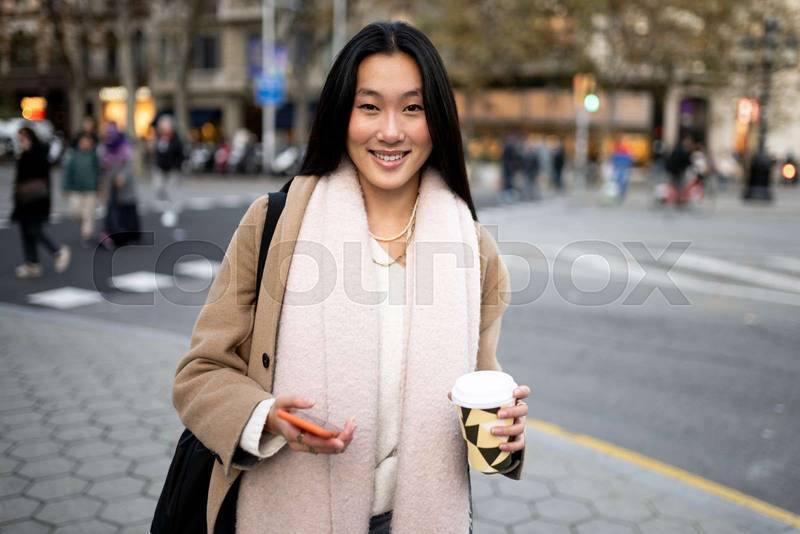 Beautiful young woman holding her phone and a cup of coffee. Satisfied confident girl smiling looking at camera in the street.