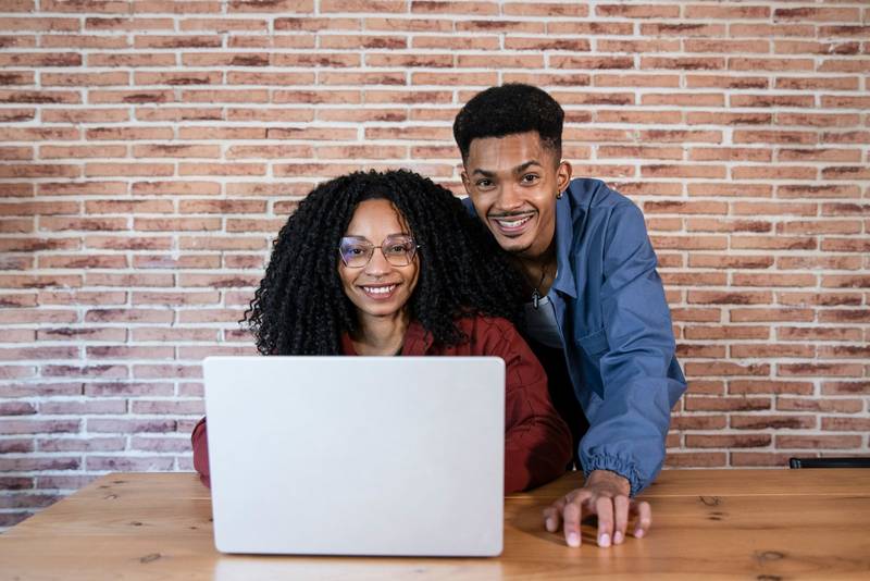 Satisfied friends looking at camera and using a computer at cozy home office. Diverse young couple using laptop together at home.