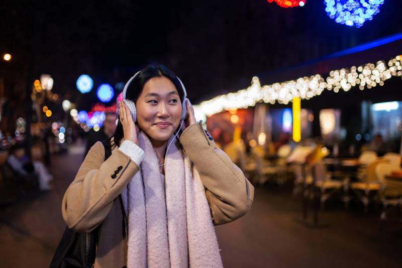 Young cheerful lady wearing headphones with christmas lights behind. Smiling girl listening to music at the street during winter.