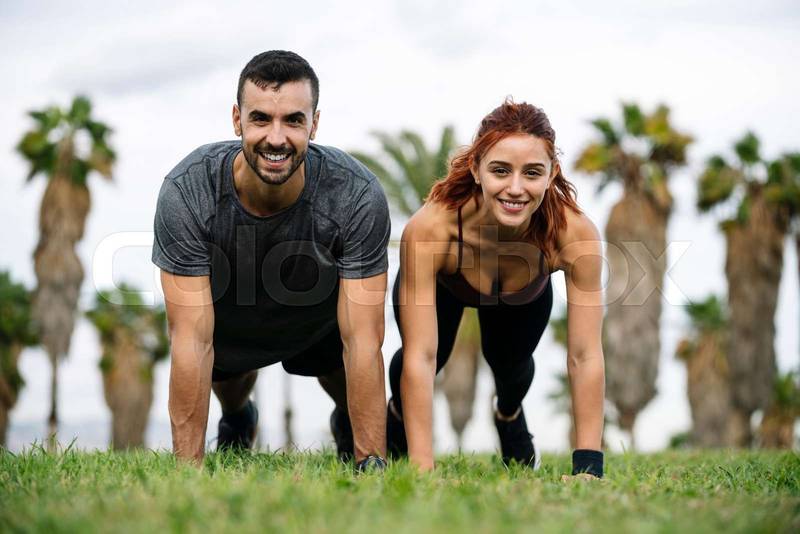 Joyful satisfied and athletic male and female sitting in the grass in sportswear training workout outside. Lovely young adult couple stretching in the floor after running together in park. 