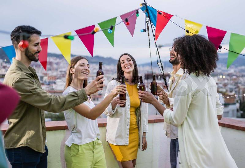 Diverse group of carefree friends having fun and drinking beer in a rooftop party in a city. Multicultural people celebrating and toasting with bottles at evening in a terrace