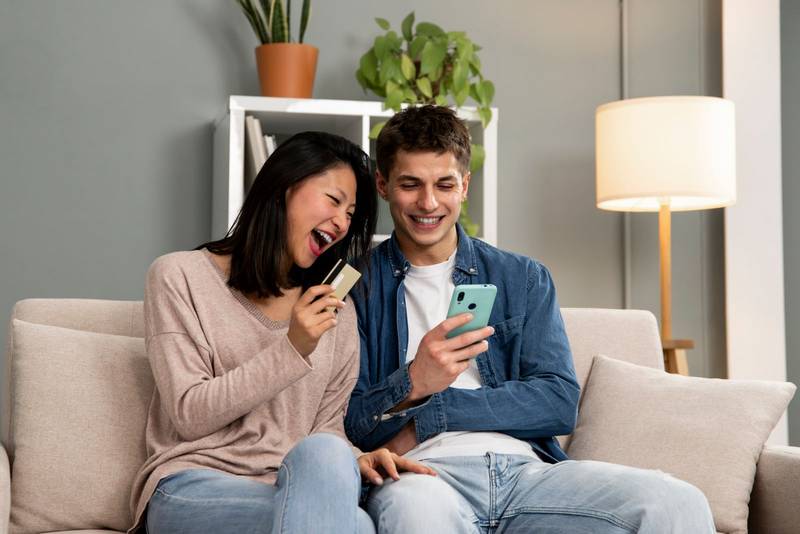 Handsome excited couple holding smartphone and credit card while sitting on a cozy sofa at home. Young adult man and woman shopping online together with phone.