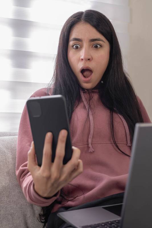 woman sitting on a sofa wearing a pink hoodie and holding a smartphone with a shocked face