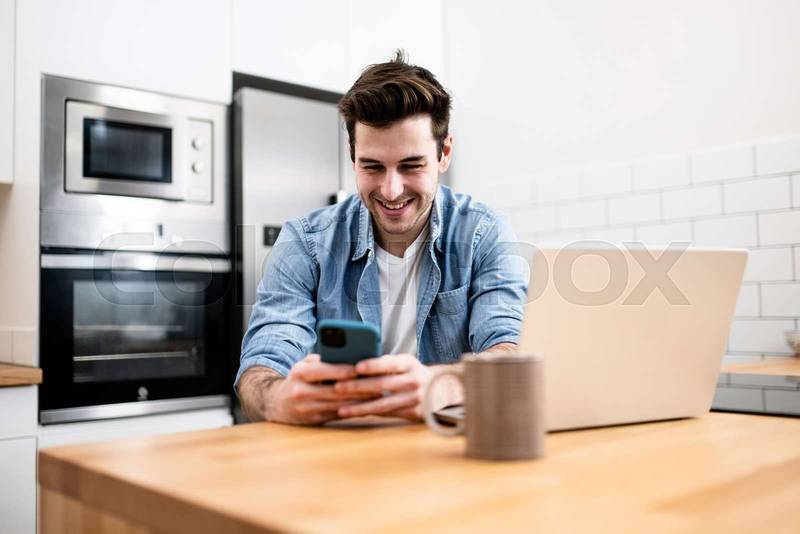 Handsome young man working with a laptop and smiling at the kitchen with a cup of tea - Cheerful, success and business concept