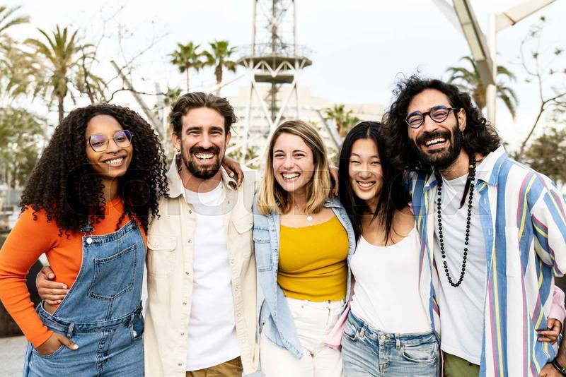 Group of cheerful millennial friends together hugging each other in street. Happy multiracial young hipster friends laughing and having fun outside looking at camera.
