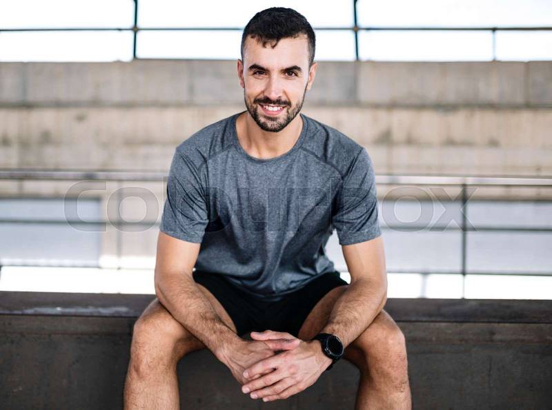 Young athlete man sitting smiling and looking at camera. Happy active runner resting satisfied from training fitness at gym.