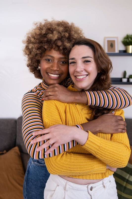 Close up of beautiful and cheerful multiracial lesbian couple hugging and looking at the camera smiling. lovely and happy homosexual women embracing at home staring at the camera.