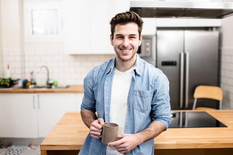 handsome happy man looking at his phone with a cup of tea in the kitchen. young man standing relaxed at home. Lifestyle concept.