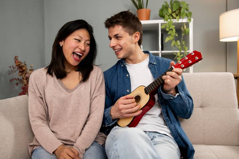Happy young diverse couple singing a song playing guitar on sofa in living room at home. Lovely young adult woman and man having fun with ukulele in their apartment.