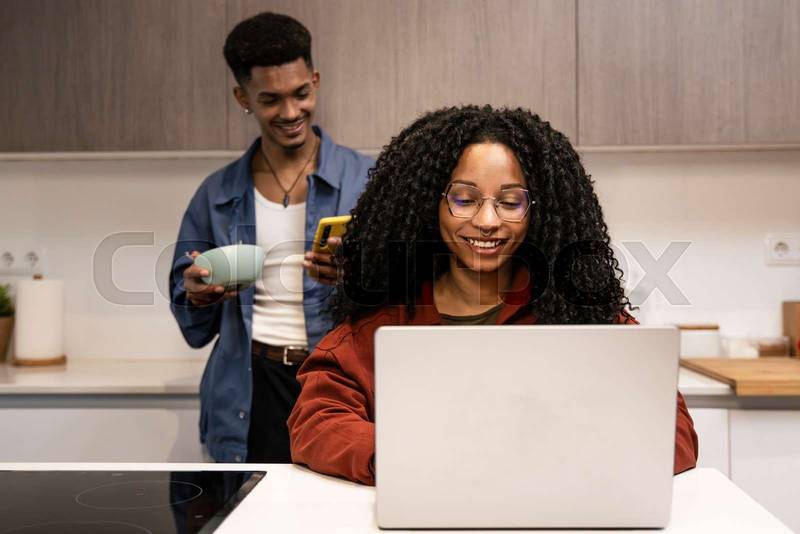 Diverse carefree couple planing together with computer and smartphone at home. Smiling man texting with phone while woman using laptop in the kitchen.