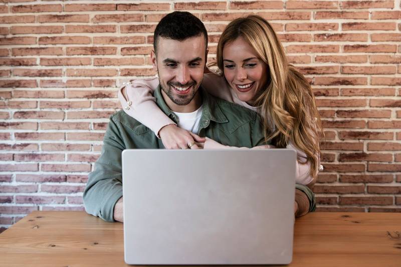 Happy couple sitting and working together from home with a laptop. Young adult man and woman using computer online in the living room.