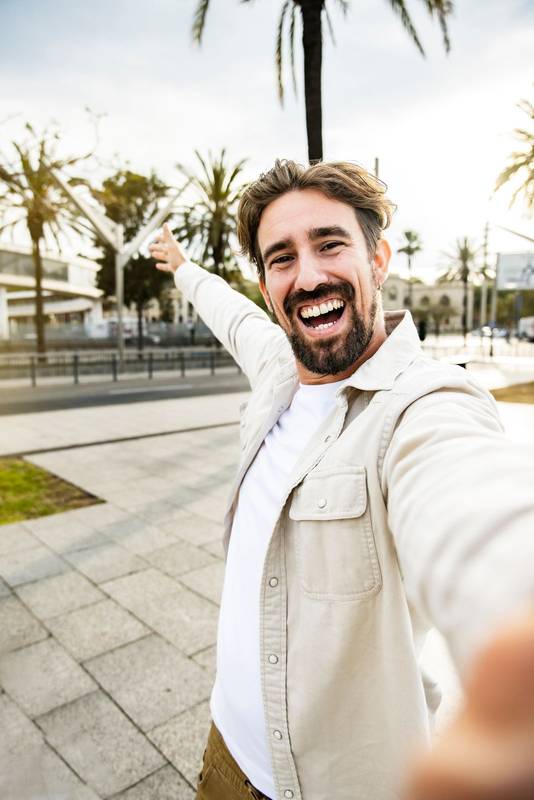 Vertical portrait of cheerful happy young man smiling looking at camera taking a selfie in the street with one arm wide open. Handsome carefree young guy laughing and staring at camera in the city.