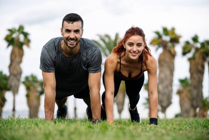 Joyful satisfied and athletic male and female sitting in the grass in sportswear training workout outside. Lovely young adult couple stretching in the floor after running together in park. 