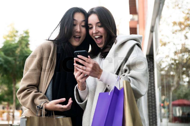 Two surprised young beautiful women holding shopping bags and looking smartphone. Cheerful friends amazed with her mobile phone. 