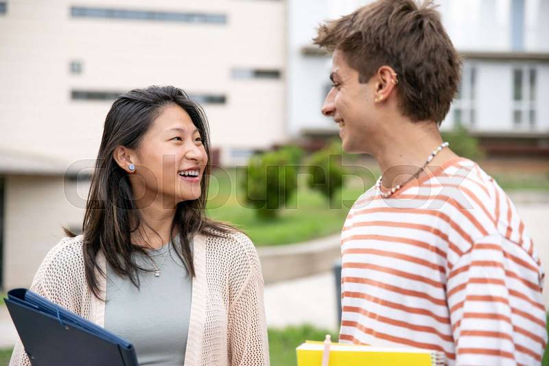 Diverse couple smiling and holding folders in a campus.Two young students looking each other smiling and satisfied in the street.