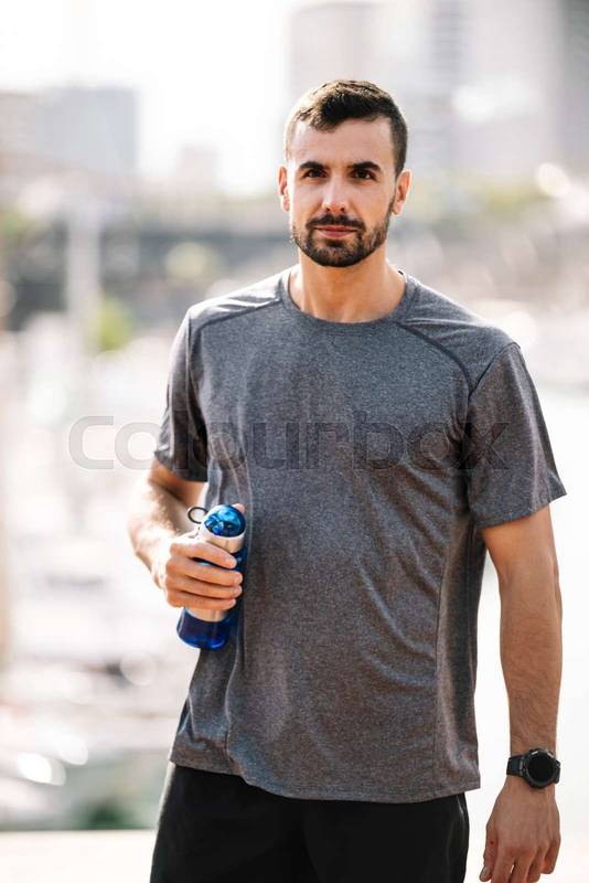 Sports fit man drinking water from bottle after training outdoor. Strong active male with recovery drink outside looking at camera.