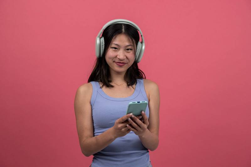 Joyful and satisfied woman with headphone texting with phone looking to the camera. Smiling young female listening to music and using online smartphone isolated on a red background.