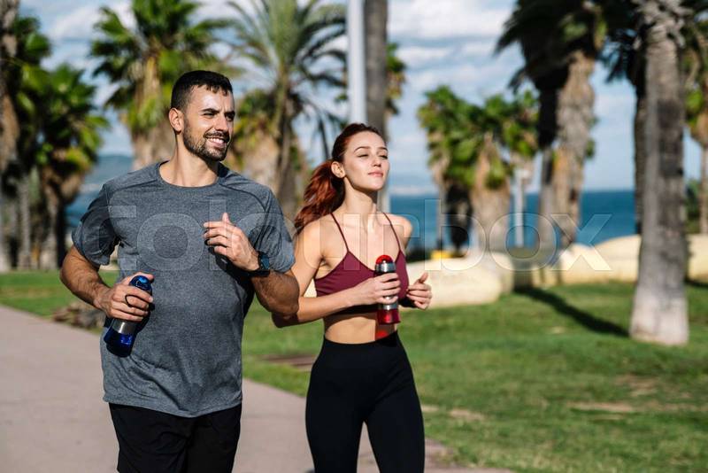 Young adult couple running together holding water bottles while jogging. Happy athletic male and female in sportswear training with drinking workout outside.