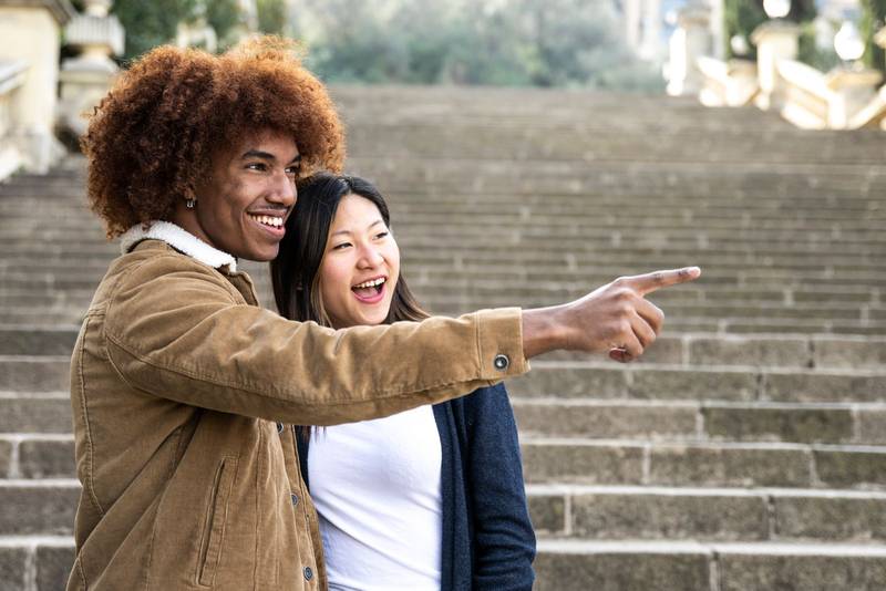 Lovely multiracial couple pointing to the right in a park.Two happy diverse young persons smiling and pointing with finger outside.