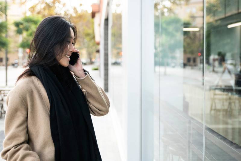 Young beautiful woman speaking smartphone and looking to showcase shop in the city. smiling shopper in front of a store