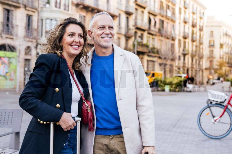 Hopeful senior man and woman standing with a suitcase happy and relaxed in old town. Mature couple hugging together with luggage in a city.