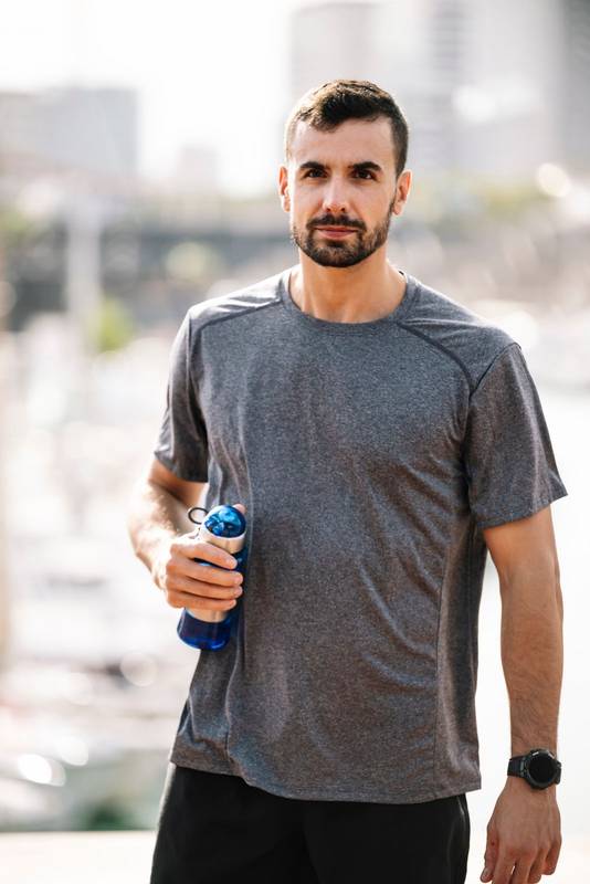 Sports fit man drinking water from bottle after training outdoor. Strong active male with recovery drink outside looking at camera.