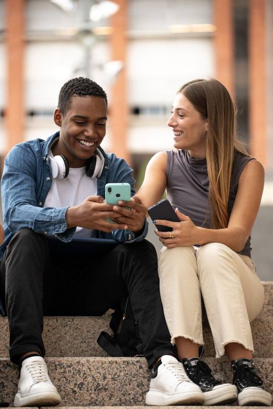 Happy young couple sitting on stairs using their phones.Two diverse friends laughing and texting outside.