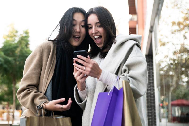 Two surprised young beautiful women holding shopping bags and looking smartphone. Cheerful friends amazed with her mobile phone. 