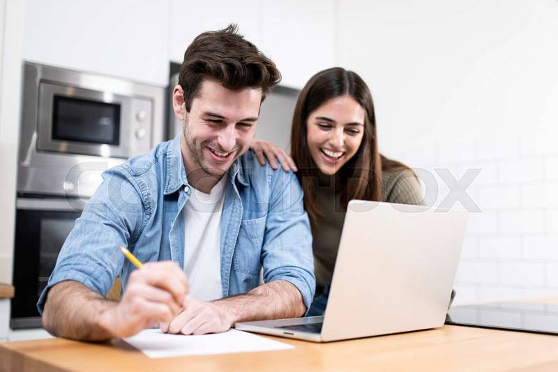young caucasian couple planning budget together at home writting notes to paper and looking a laptop. two cheerful young adults sitting on the kitchen. finance concept