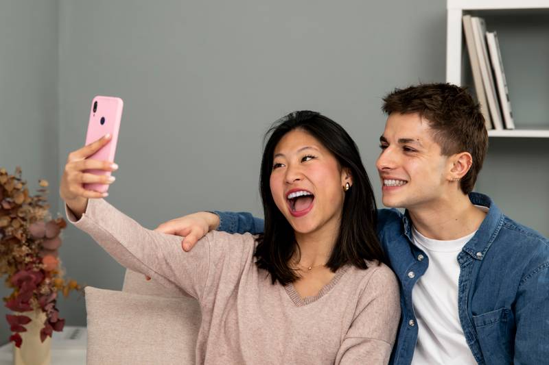 Cheerful man and woman laughing in a video call with smart phone. Diverse young couple on the sofa in their living room taking a selfie waving. 