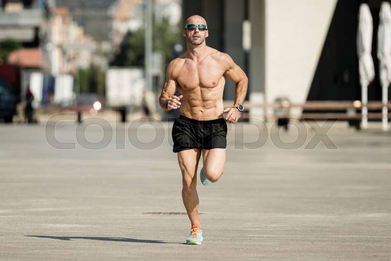 Athletic young man running in the street wearing sunglasses. Front view of sportsman jogging outdoors shirtless.