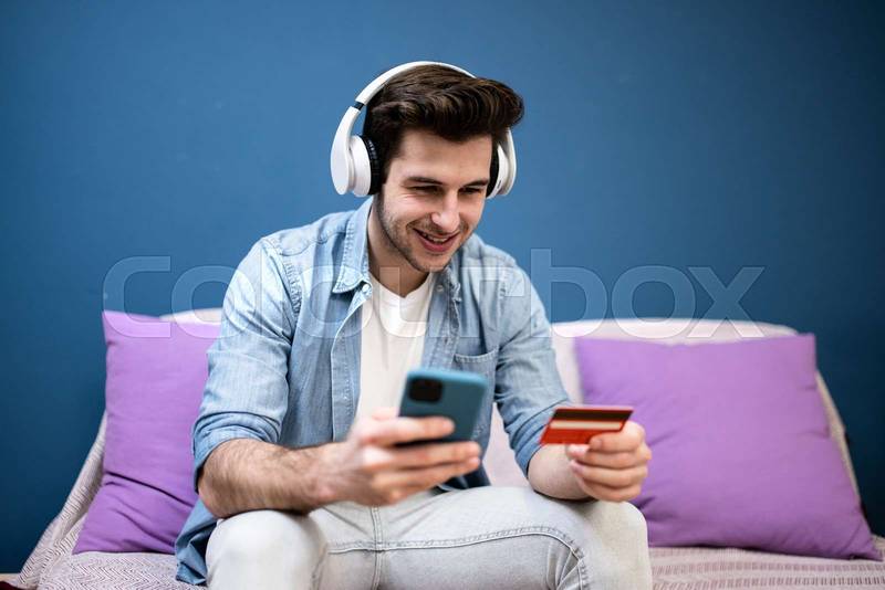 Man sitting in front of the computer holding a credit card at home - Young adult doing a online payment with his laptop at kitchen - business, technology concept