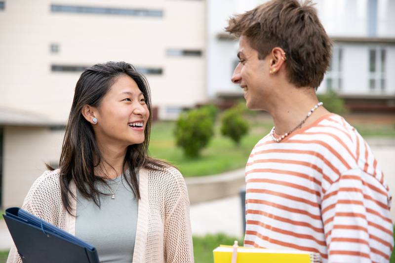 Diverse couple smiling and holding folders in a campus.Two young students looking each other smiling and satisfied in the street.