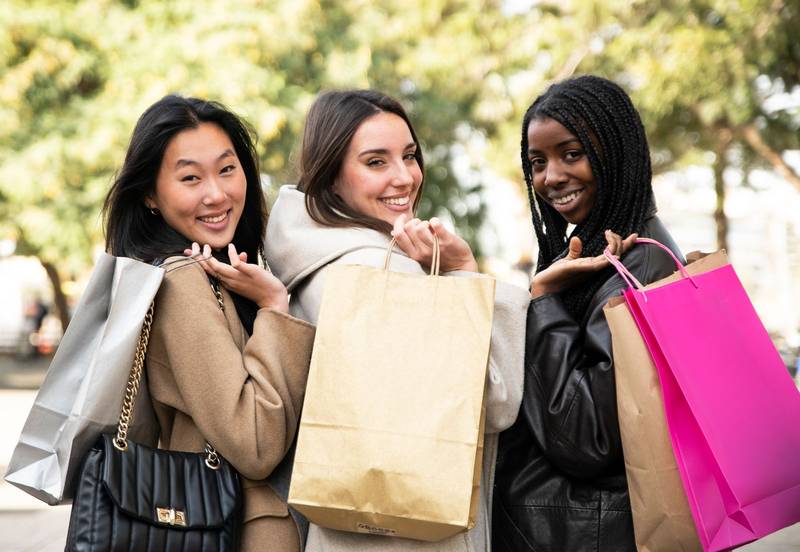Three cheerful young women shopping in the city - Rear view of three happy beautiful young women with shopping bags in the city looking at camera - Fashion and friendship concept