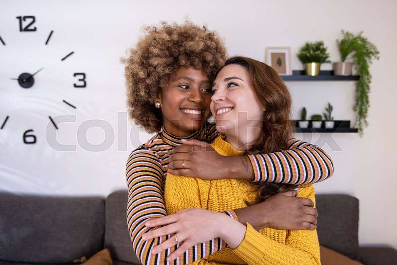 Beautiful and cheerful multiracial lesbian couple hugging and looking each other smiling. Lovely and happy homosexual women embracing at home staring each other.