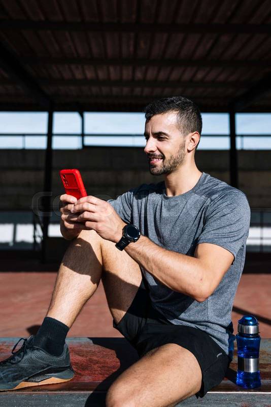 Fit man using smartphone relax resting from exercise in a bench outside. Active sports athlete texting with phone during training.