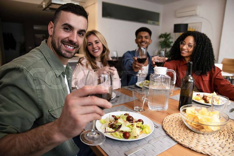 Cheerful diverse group of happy friends having party diner at home. Smiling man toasting with people eating and drinking on a modern apartment. 