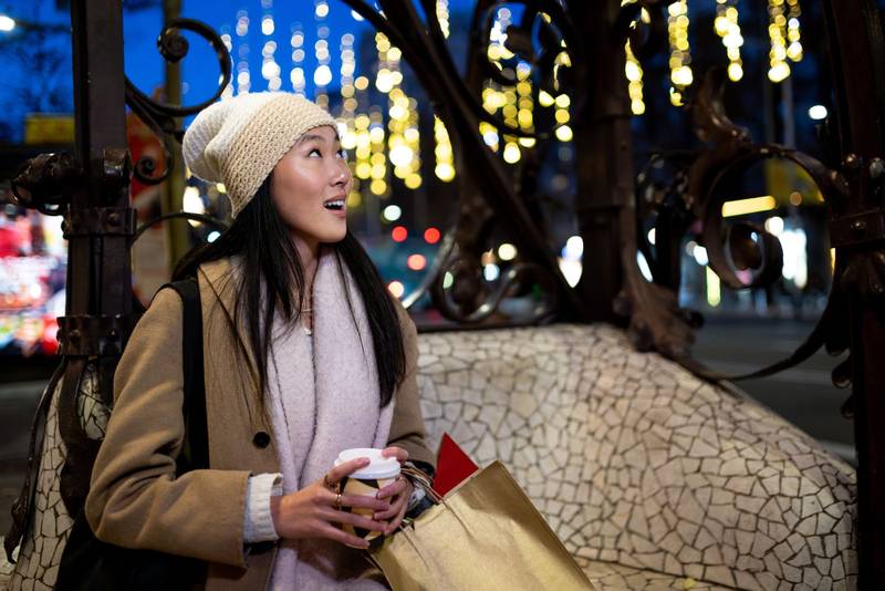 Joyful young woman shopping at evening staring at beautiful lights. Expressive female holding a coffee looking up at night in the street.