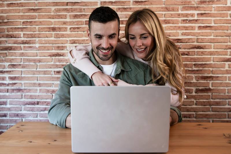 Happy couple sitting and working together from home with a laptop. Young adult man and woman using computer online in the living room.