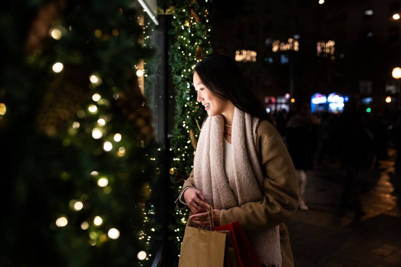 Joyful young woman shopping at evening staring at shop window. Smiling female holding bags at night in the street.