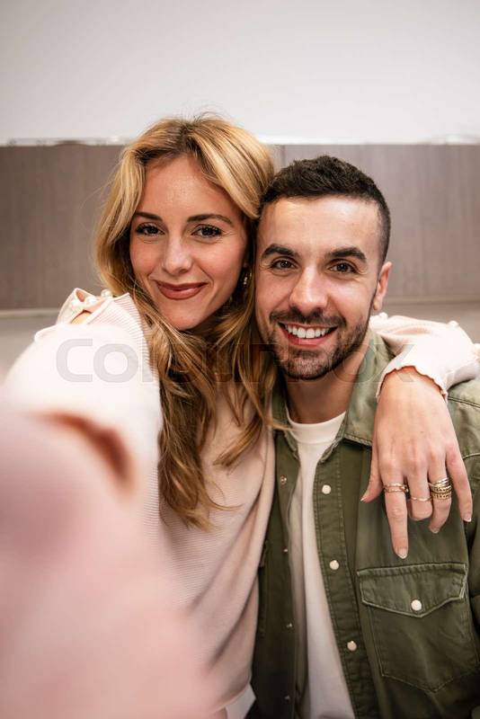Carefree man and woman relaxing together taking a selfie at living room. Affectionate smiling young adult couple embracing at home looking at camera. 