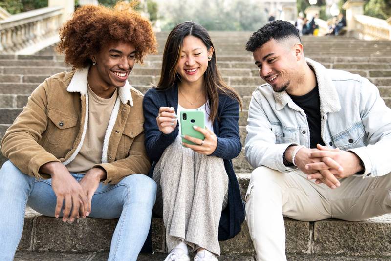 Three young diverse people looking at a smartphone sitting on the stairs.Two young men smiling and watching at the phone of a happy woman.
