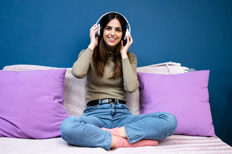 Cheerful young woman listening to music and smiling sitting on couch in livinig room. Happy young girl having fun at home.