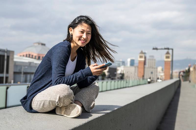 Young beautiful girl sitting on bank relaxed and texting with her phone looking at camera. Satisfied woman using an smartphone sitting in the street staring at camera.