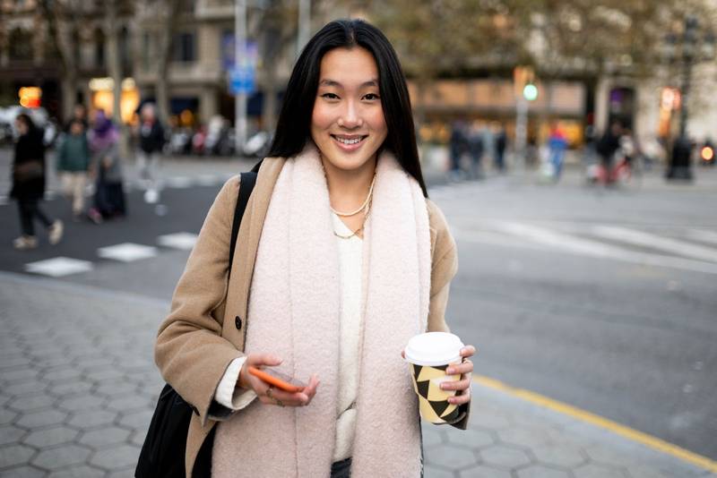 Beautiful young woman holding her phone and a cup of coffee. Satisfied confident girl smiling looking at camera in the street.