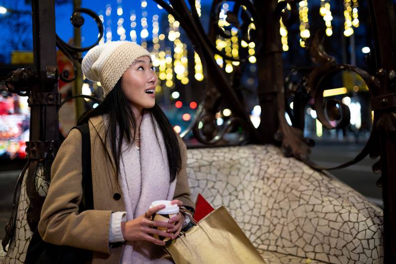 Joyful young woman shopping at evening staring at beautiful lights. Expressive female holding a coffee looking up at night in the street.