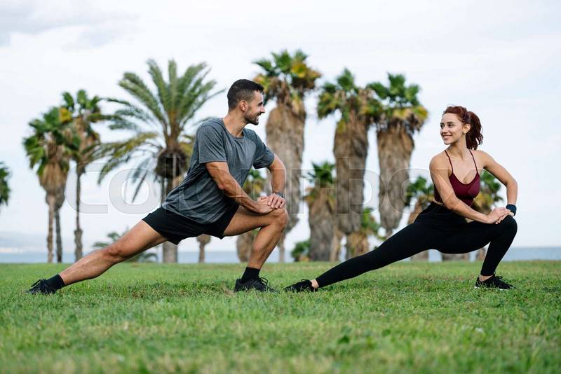 Joyful satisfied and athletic male and female sitting in the grass in sportswear training workout outside. Lovely young adult couple stretching in the floor after running together in park. 