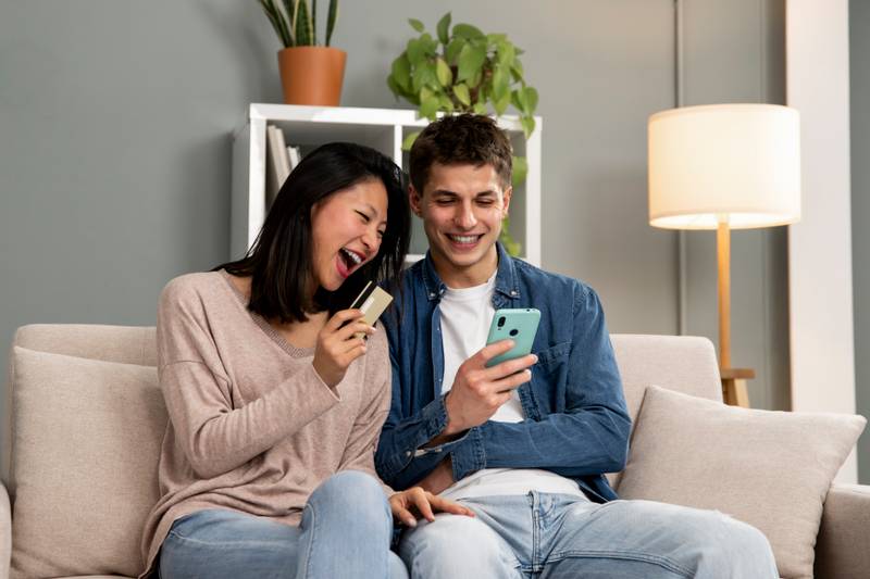 Handsome excited couple holding smartphone and credit card while sitting on a cozy sofa at home. Young adult man and woman shopping online together with phone.